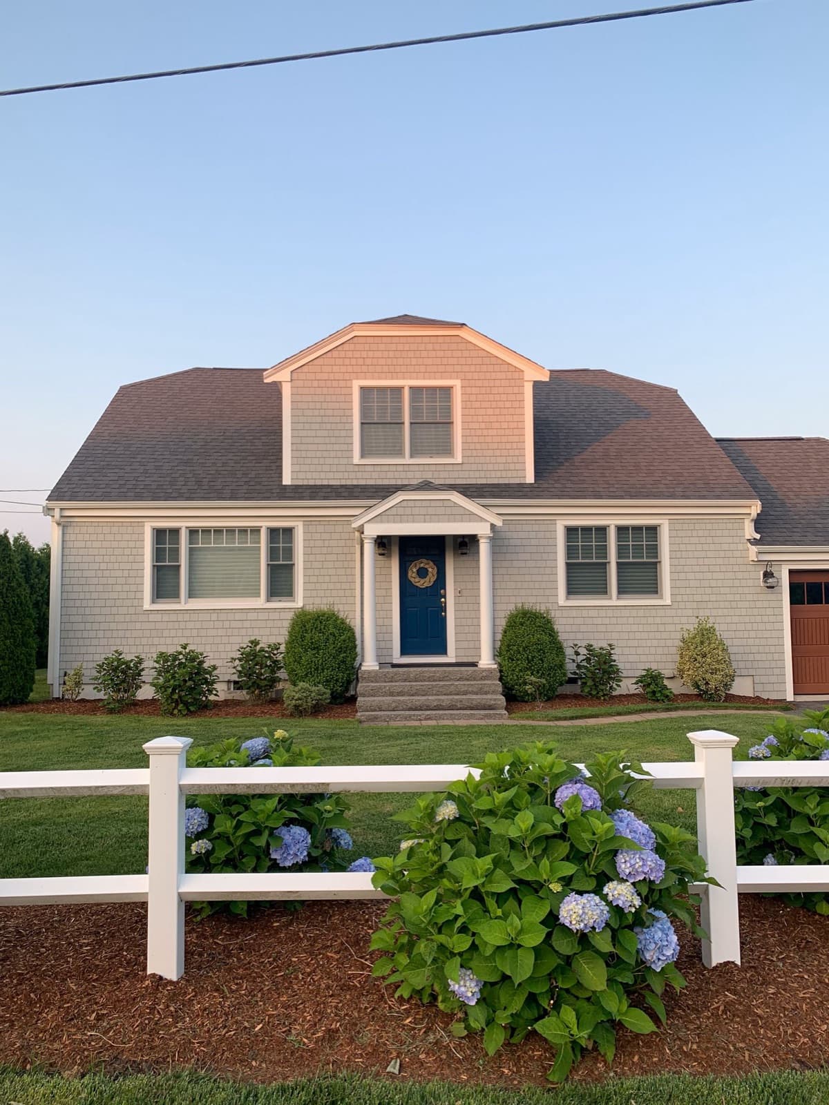 Gray-shingled Cape Cod style house with a blue front door and blooming hydrangeas